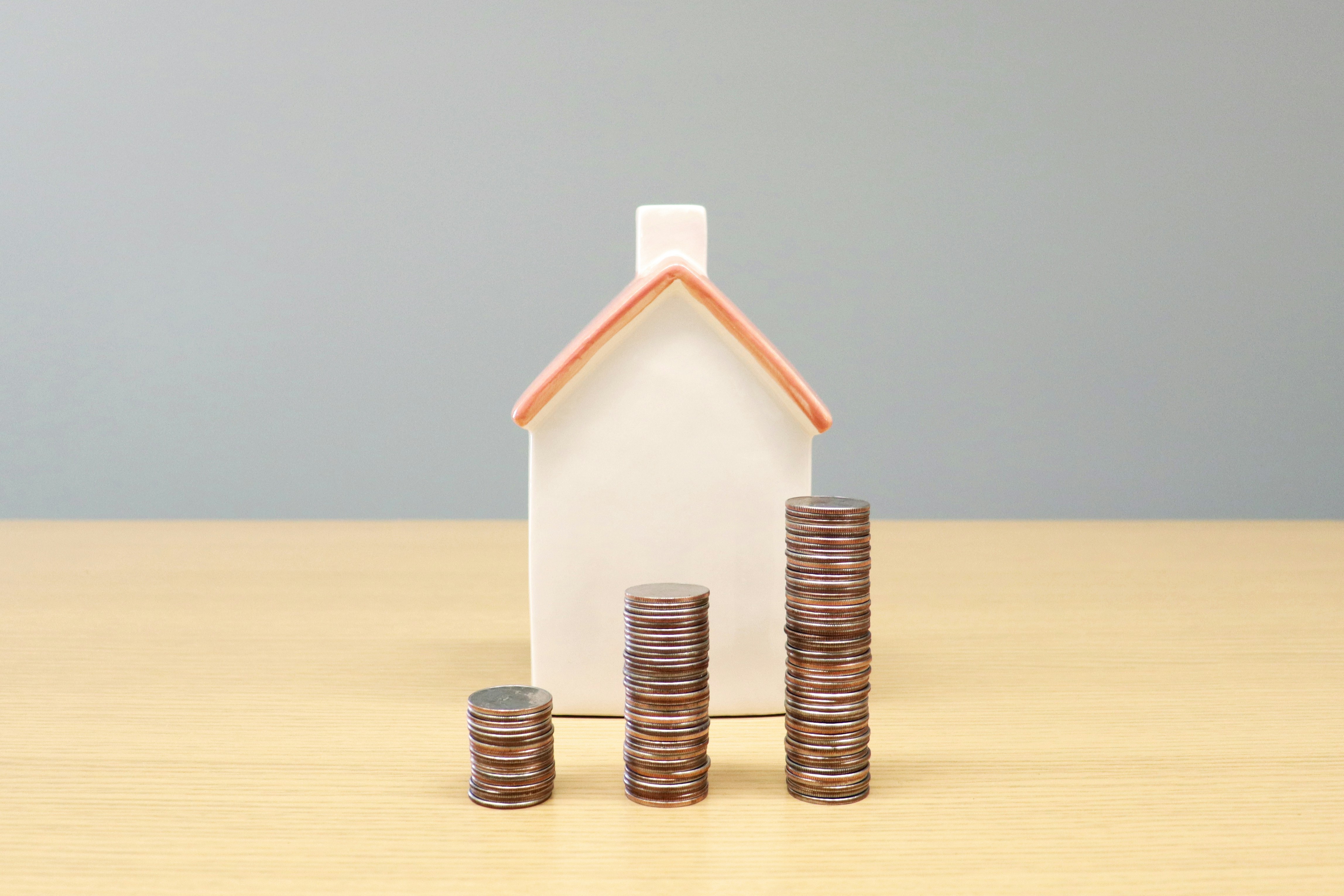 Small white house figurine behind three ascending stacks of coins on a wooden table.