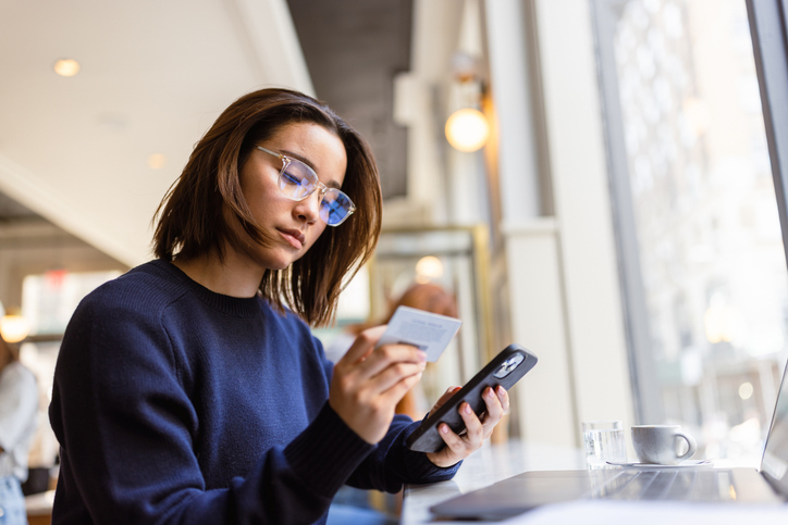 A focused woman wearing glasses is holding a credit card and looking at her phone, possibly managing her finances or making a payment in a cafe setting.